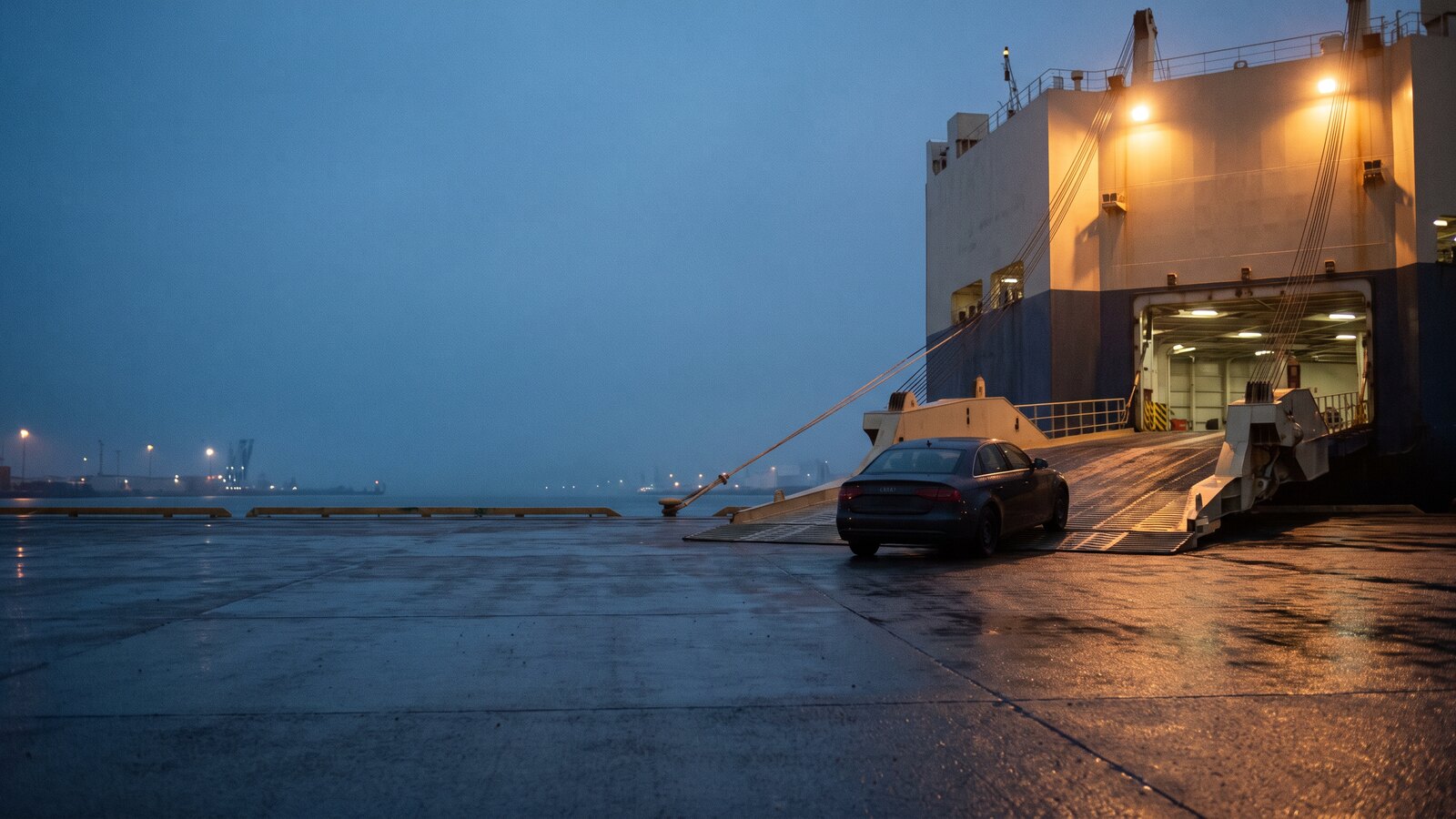 A car waiting at the foot of a floodlit stern ramp leading into a vehicle carrier on a wet quayside at dusk.